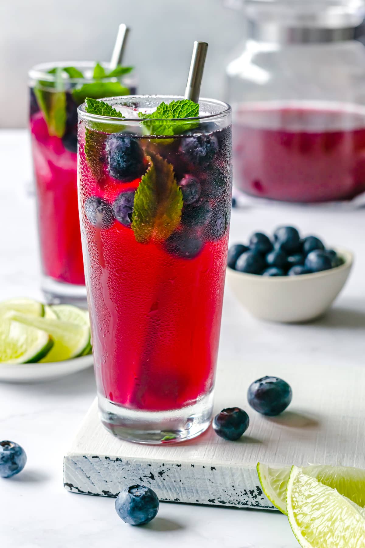 Blueberry juice with fresh blueberries and mint, in a clear tall cup with silver straw on a white wood board with lim slices around and blueberries.