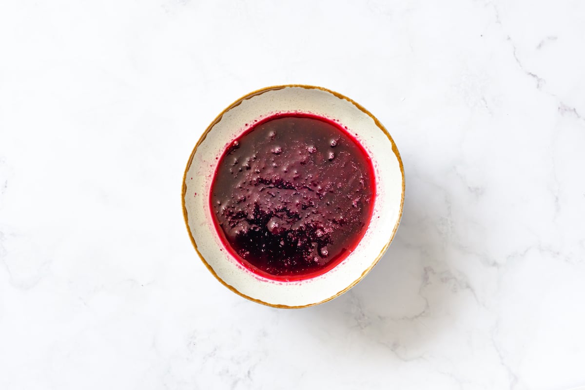 Meshed and boiled blueberries, mint, honey, and lime zest in a cream and brown shallow bowl on white marble background.