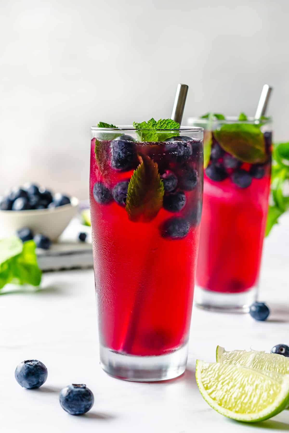 two fresh made blueberry refresher with blueberries and mint in clear tall glass cups, slices of lime and blueberries around on white background.