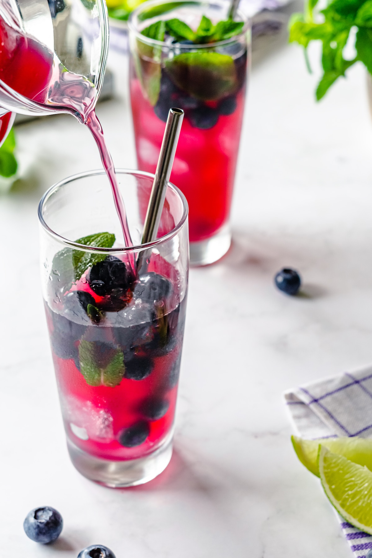 Blueberry refresher being poured into a clear tall cup with fresh blueberries and mint and a silver straw.