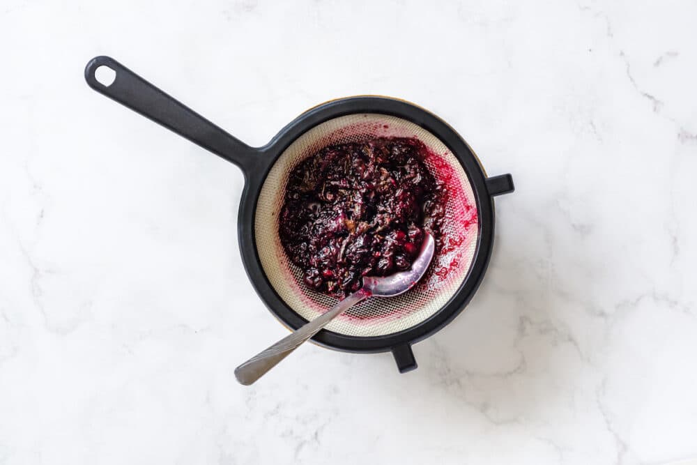 fine black mesh on top of white and brown bowl, silver tablespoon meshing through boiled blueberries, mint, lime zest, juice.