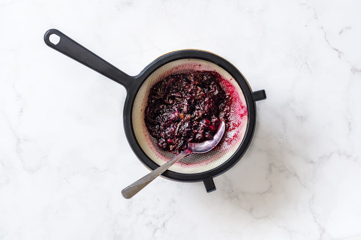 Fine black mesh on top of white and brown bowl, silver tablespoon meshing through boiled blueberries, mint, lime zest, juice.