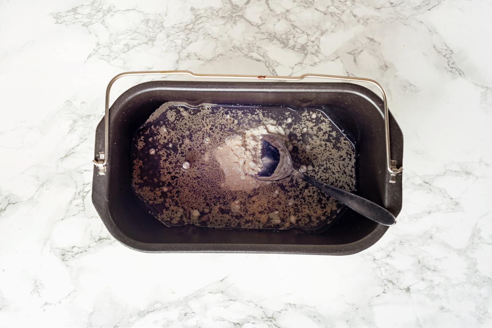 Bread ingredients in a bread machine bucket mixing with a spoon.