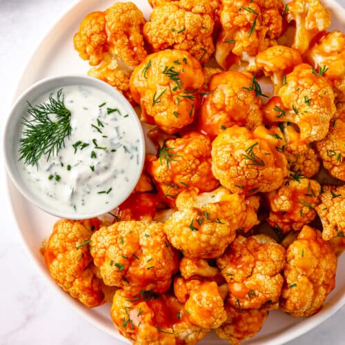 on a white plate, buffalo baked cauliflower wings with fresh dill, and a side of greek yogurt dipping sauce.