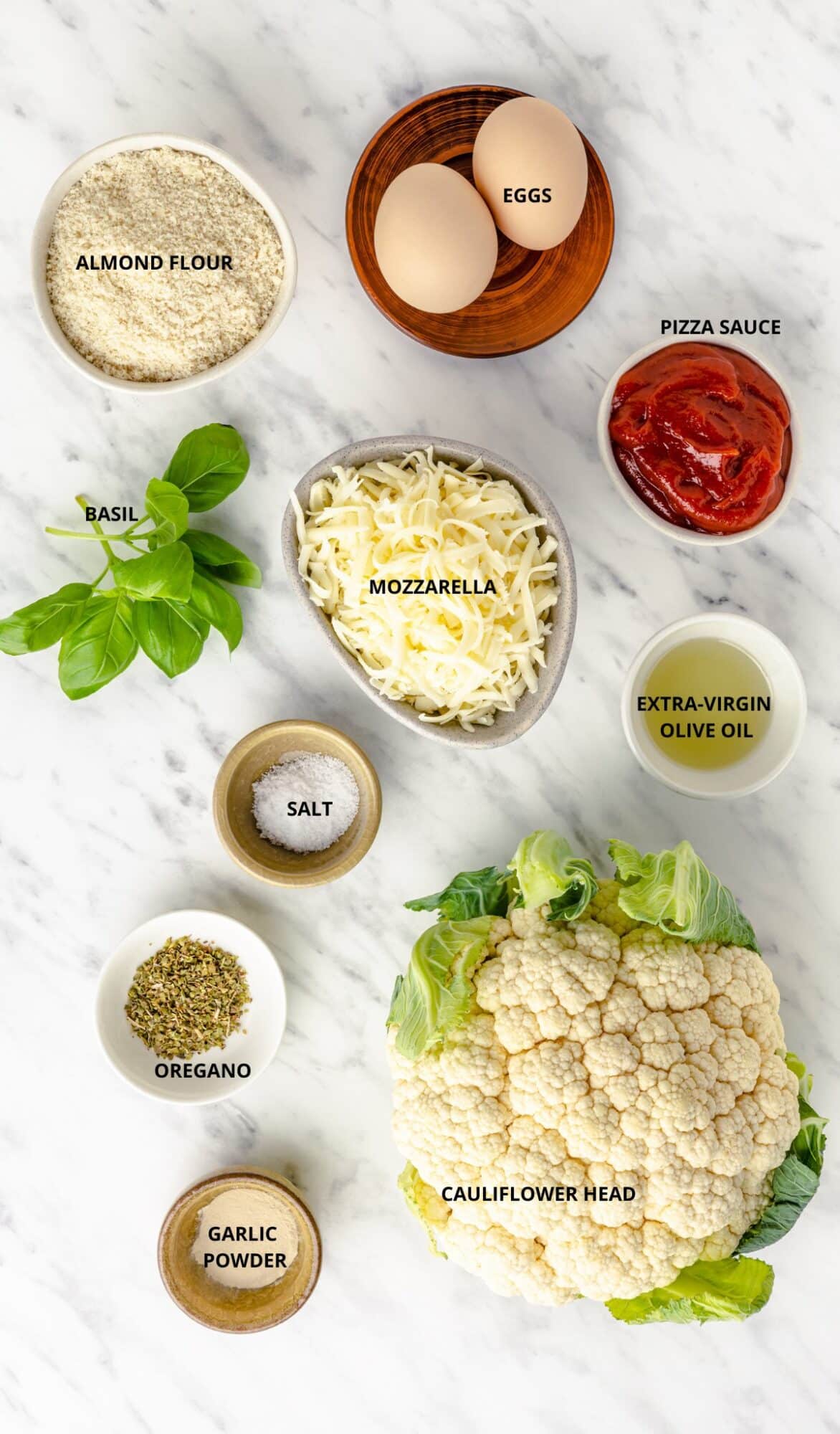 cauliflower pizza ingredients, cauliflower head, mozzarella grated, fresh basil, pizza sauce, oregano dried, salt, olive oil, almond flour and two eggs all individually laid out.