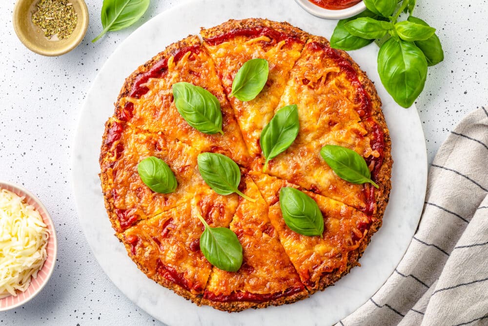 cauliflower crust pizza baked with pizza sauce and grated cheese with basil on top on a marble background.