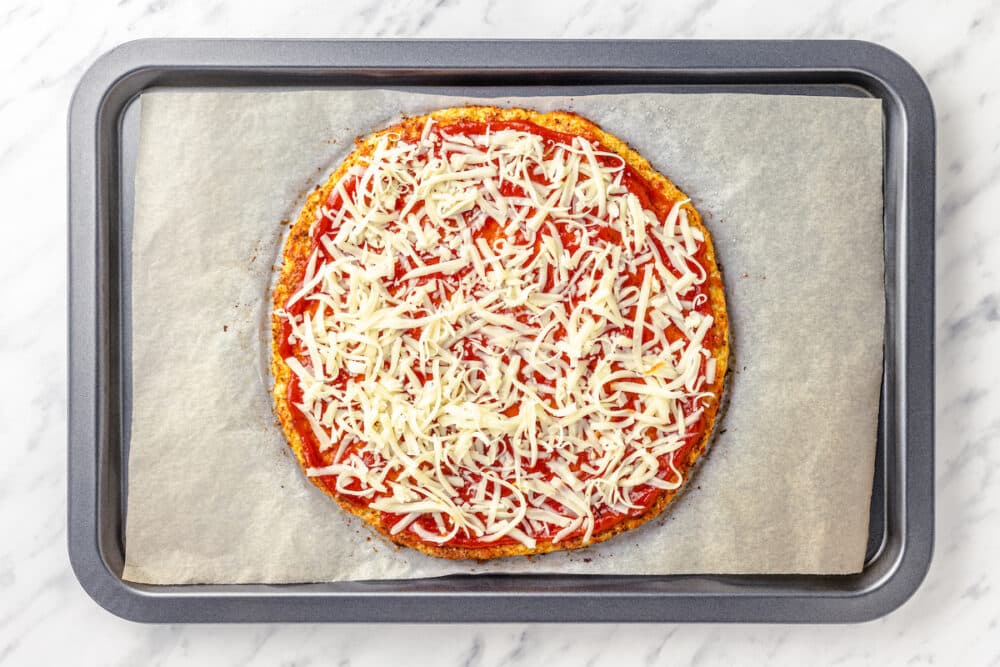 baked cauliflower crust with pizza red sauce and grated mozzarella on top on a baking sheet.