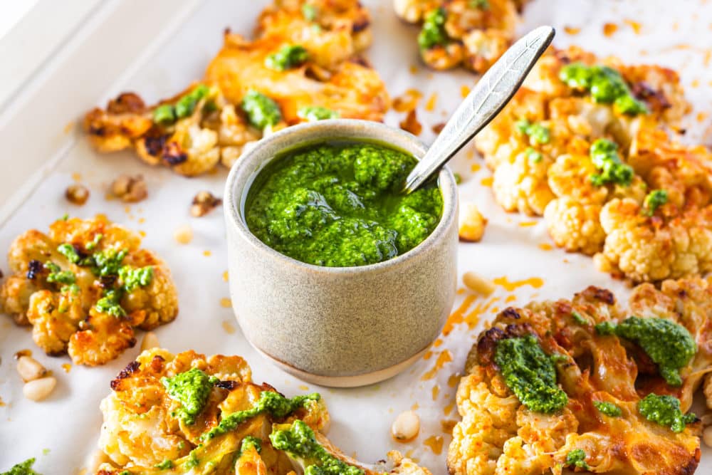 a baking sheet with a bowl of pesto sauce and a spoon with baked cauliflower steak.