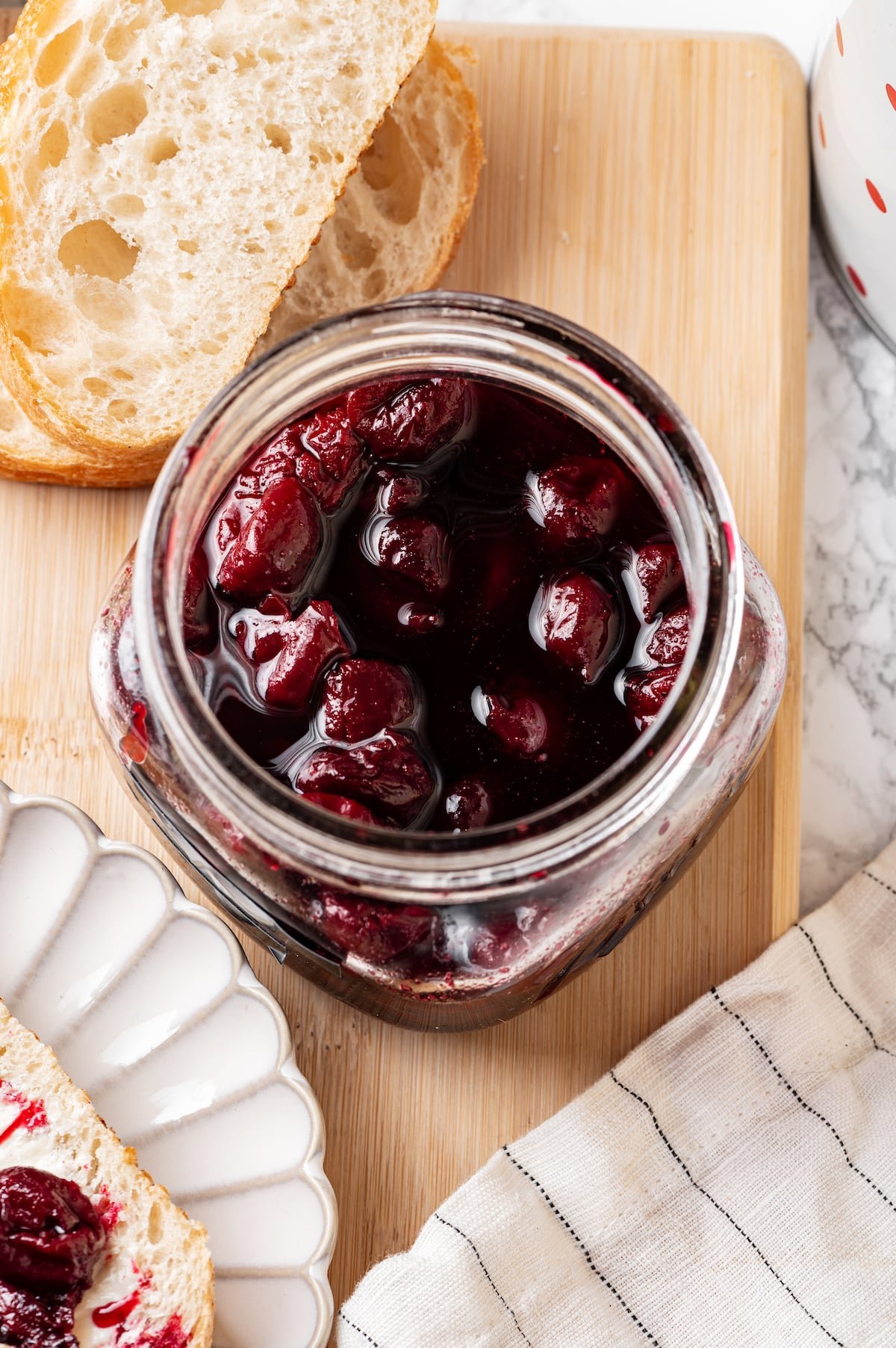 red cherry jam on a wooden board with slices of white bread on the side and beige towel with black stripes on another side.