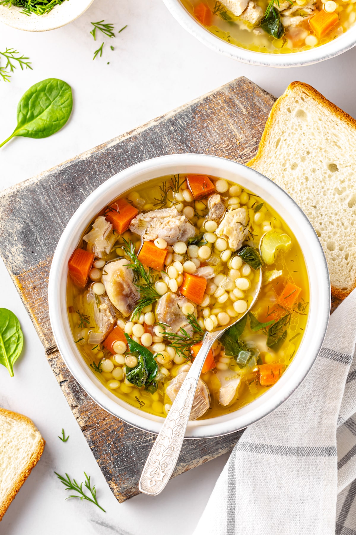 soup with couscous and vegetables in a white bowl on top of a wooden board with a towel on the side and a slice of bread on the wooden board next to the white bowl with a spoon and the couscous soup in it.