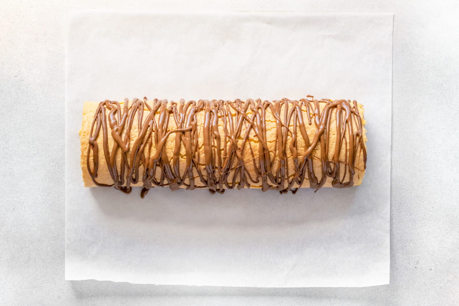 Chocolate drizzled atop strawberry cake roll on parchment paper.