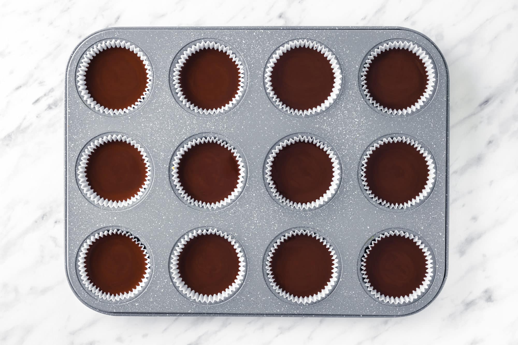 A grey muffin tin with white muffin cups and chocolate in them.