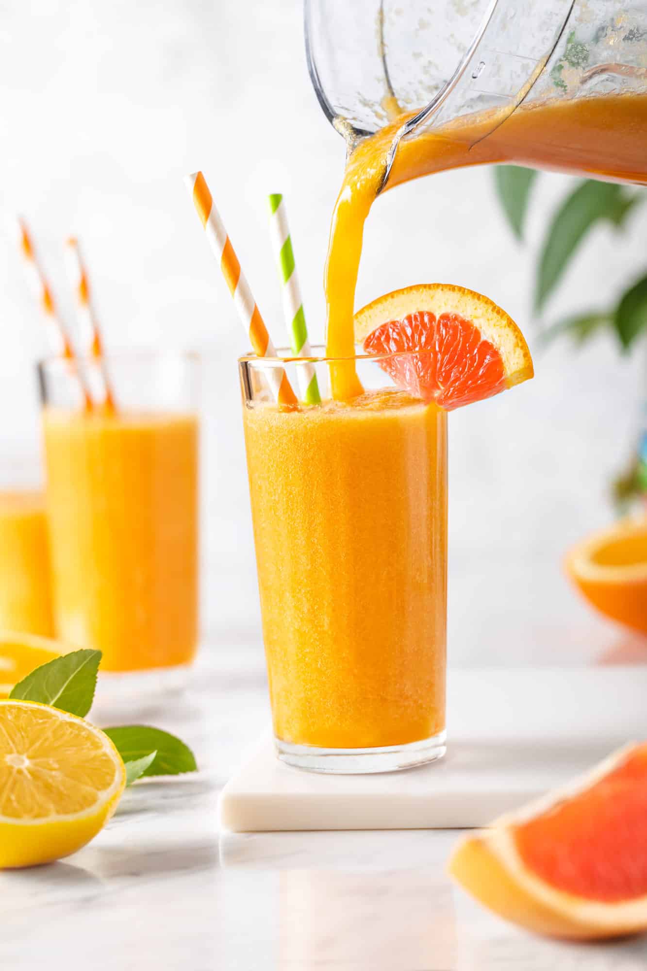 Citrus smoothie being poured into a glass in a glass with an orange wedge on the rim and two straws.