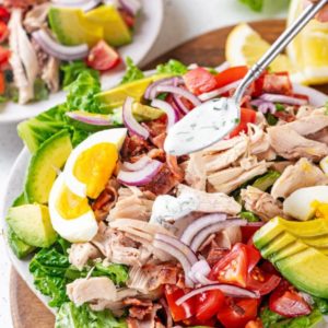 Cobb salad with chicken, tomatoes, and hard boiled eggs on a bed of lettuce on a white plate, with ranch drizzling on top.