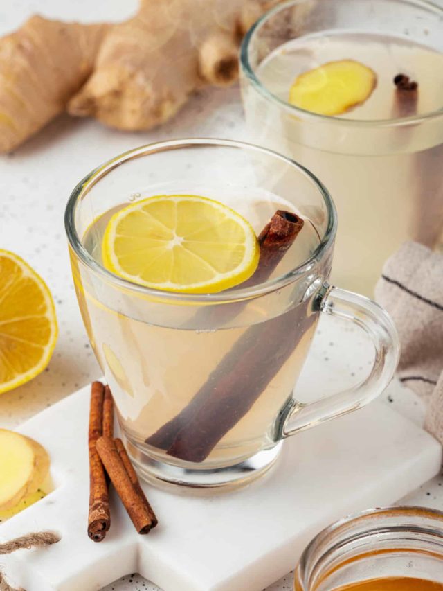 light clear ginger tea in a glass mug with a cinnamon stick and lemon slice in the mug on a white board that has cinnamon stick on it too.