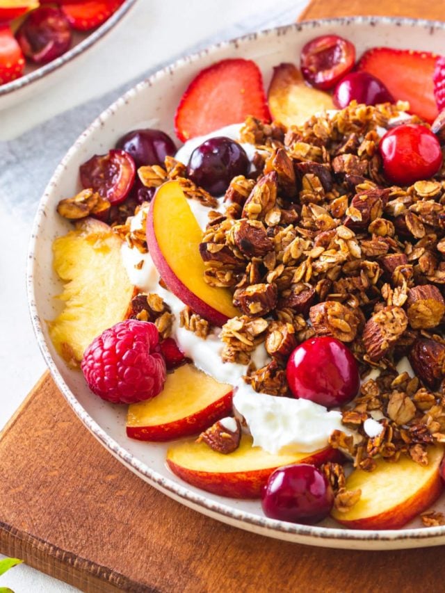 A bowl of muesli with yogurt and fruit on a wooden cutting board.