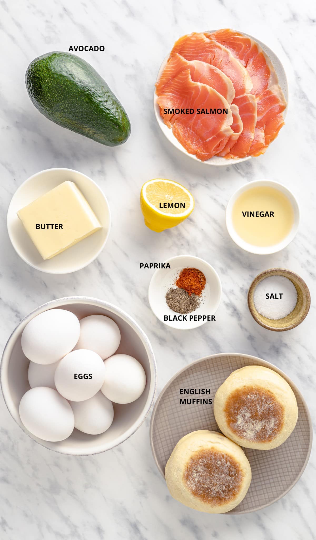 ingredients to make eggs benedict with salmon including smoked salmon, avocado, lemon, butter, vinegar, paprika, black pepper, salt, eggs, and english muffins. 