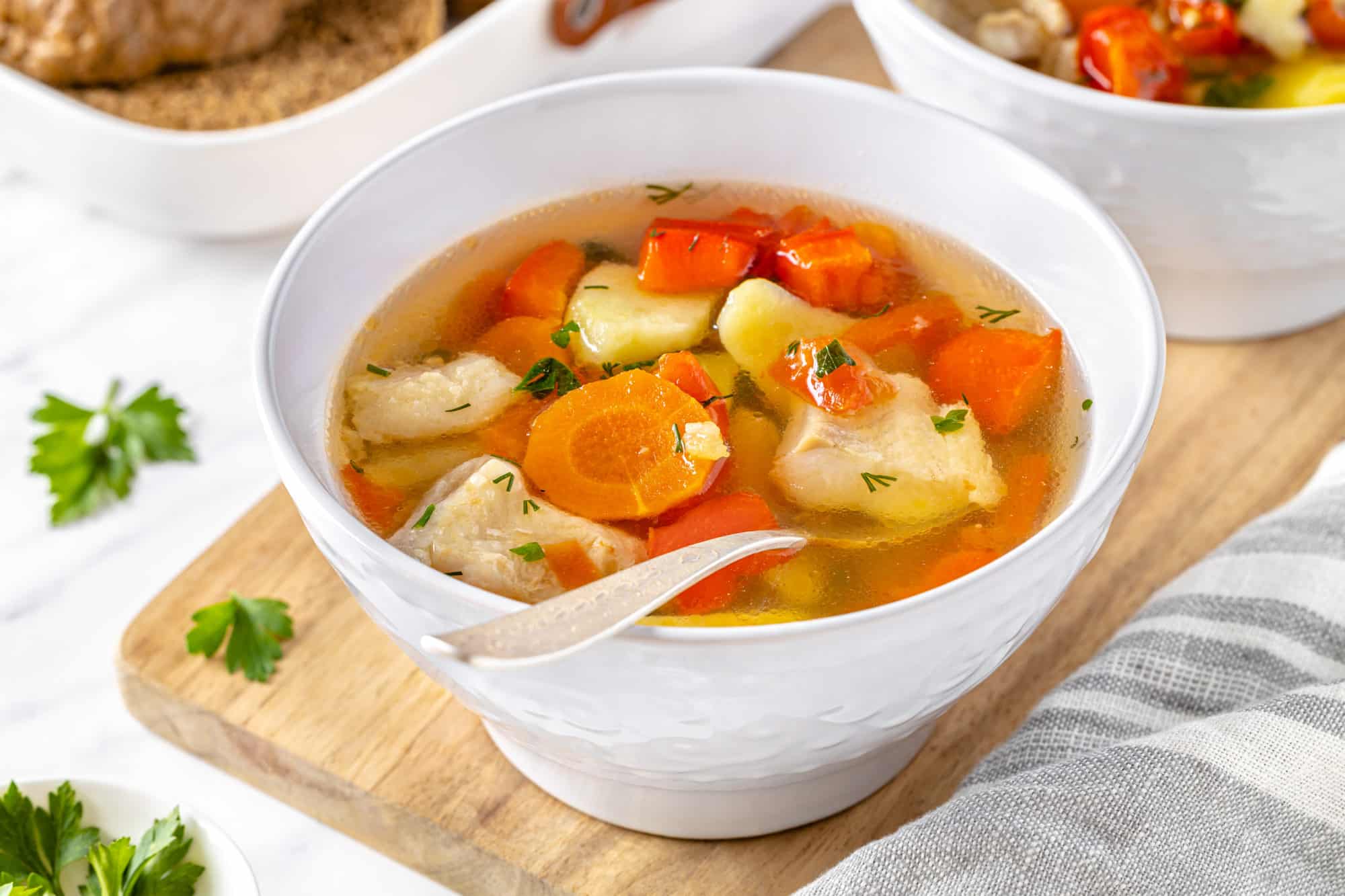 a white bowl of fish soup with veggies floating in it on a wooden board with a spoon in the soup.