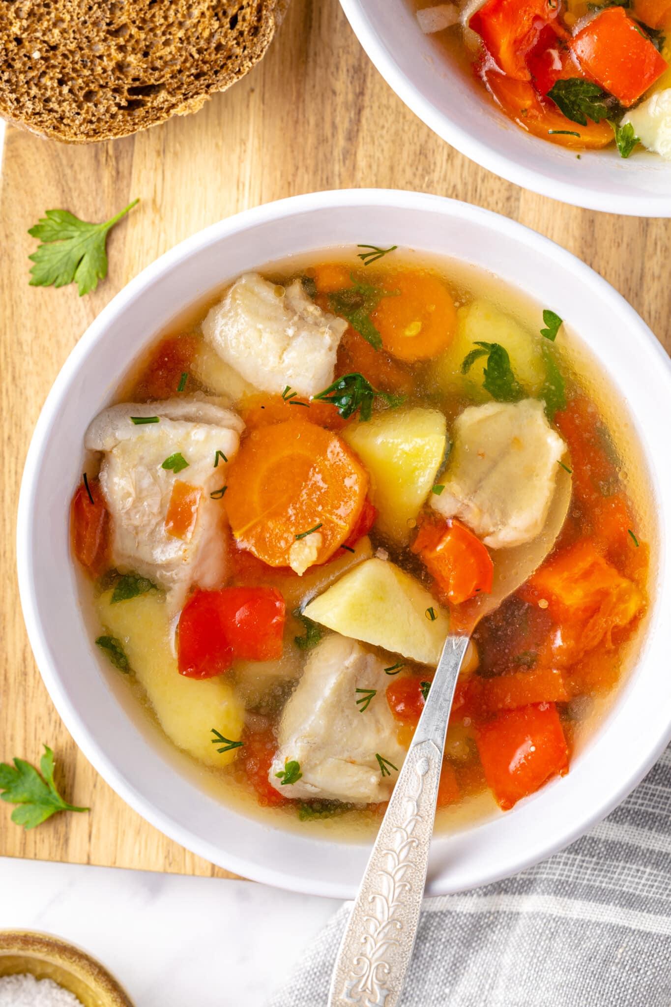 a wooden board with a white bowl on it filled with a fish soup and a spoon with a slice of bread and parsley on the side.