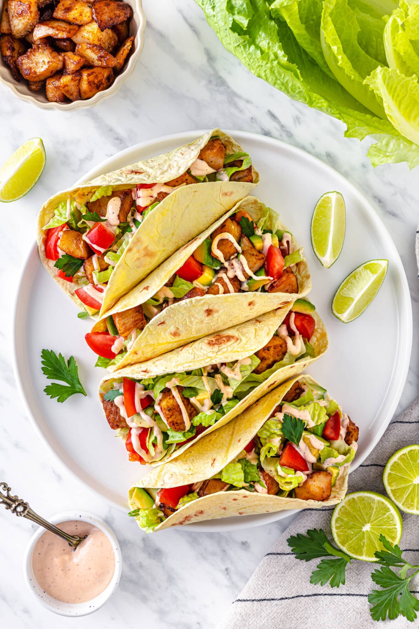 Four fish tacos all together with drizzled spicy sauce and lime wedges on the side.