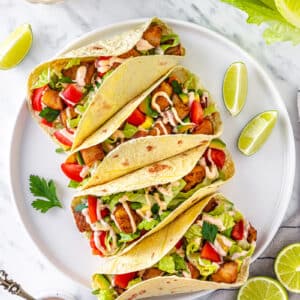 tacos with seafood and vegetable filling on a white plate with limes and other ingredients around.