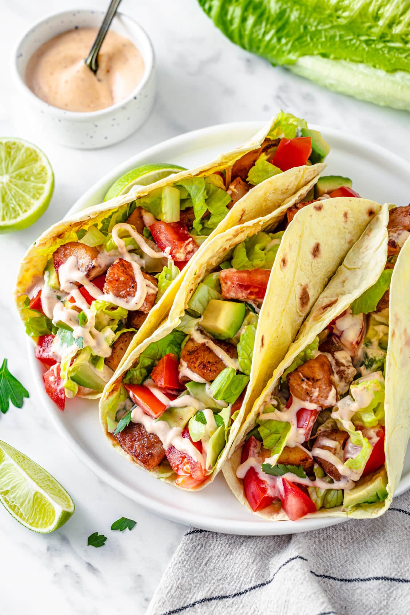 Soft taco shells filled with fish, chopped veggies, and drizzled with spicy fish sauce with lime wedges on the side for serving.