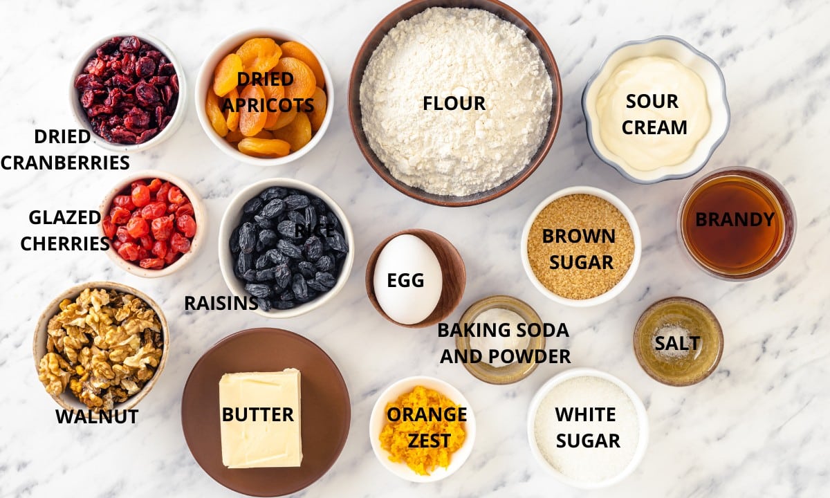 fruit cake ingredients in a different bowls on a white background flour baking soda baking powder salt sour cream butter egg white sugar brown sugar orange zest walnuts raisins dried apricots cherries dried cranberries and brandy.