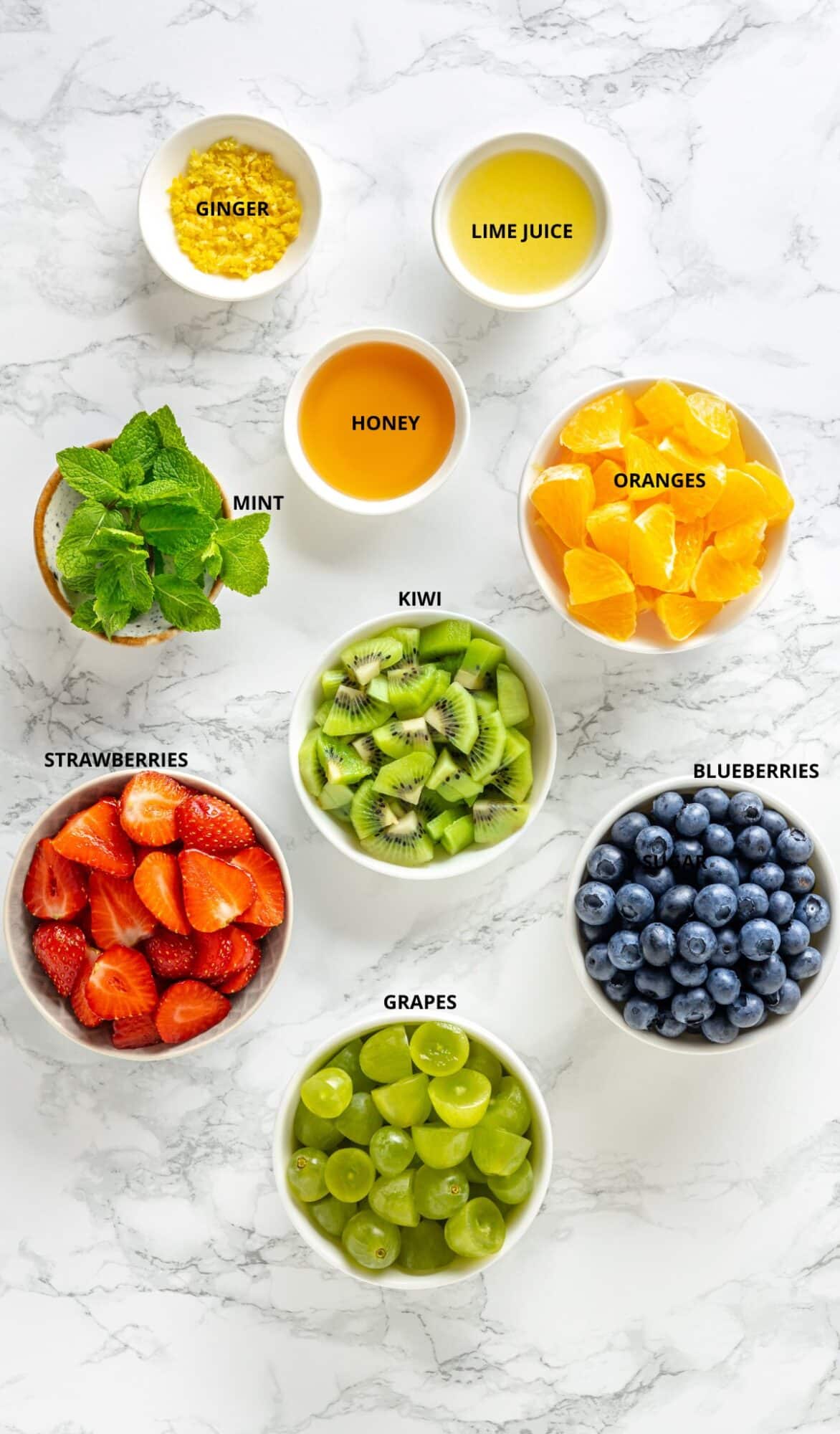 marble background, with individual bowls with different fruit, blueberries, strawberries, oranges, mint, honey, kiwis, grapes, ginger grated and lime juice.