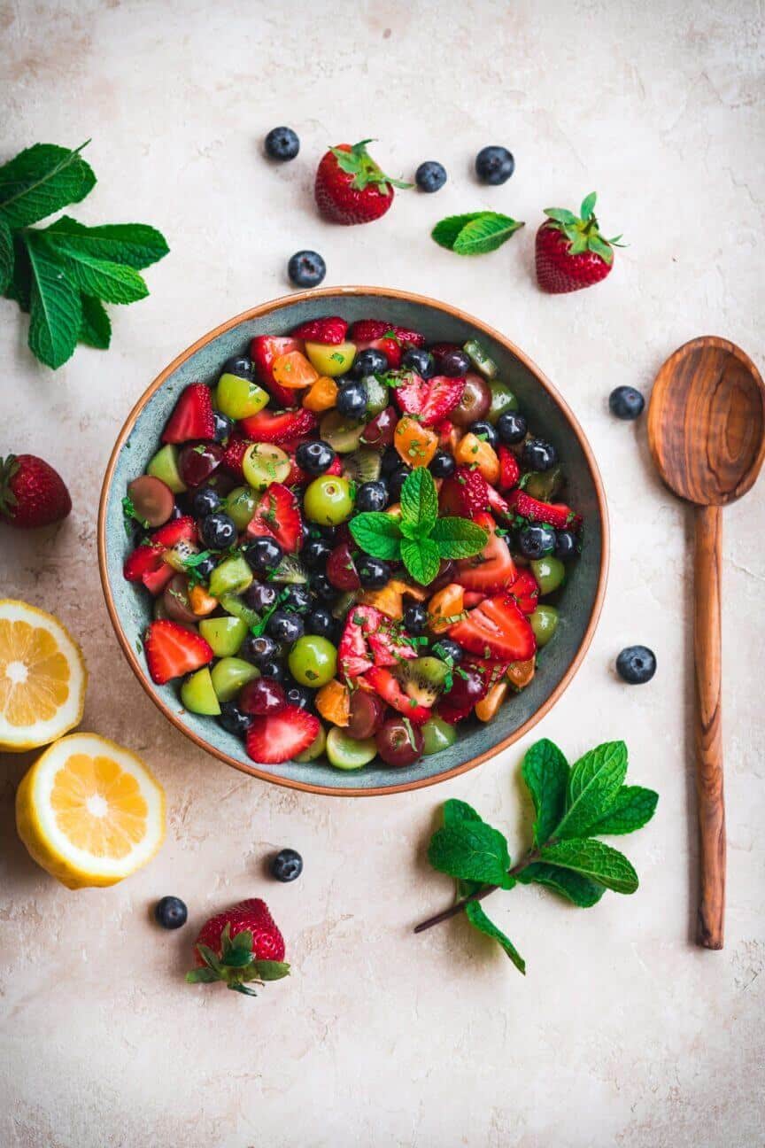 Fruit salad in a bowl with a wooden spoon and fresh fruit around.