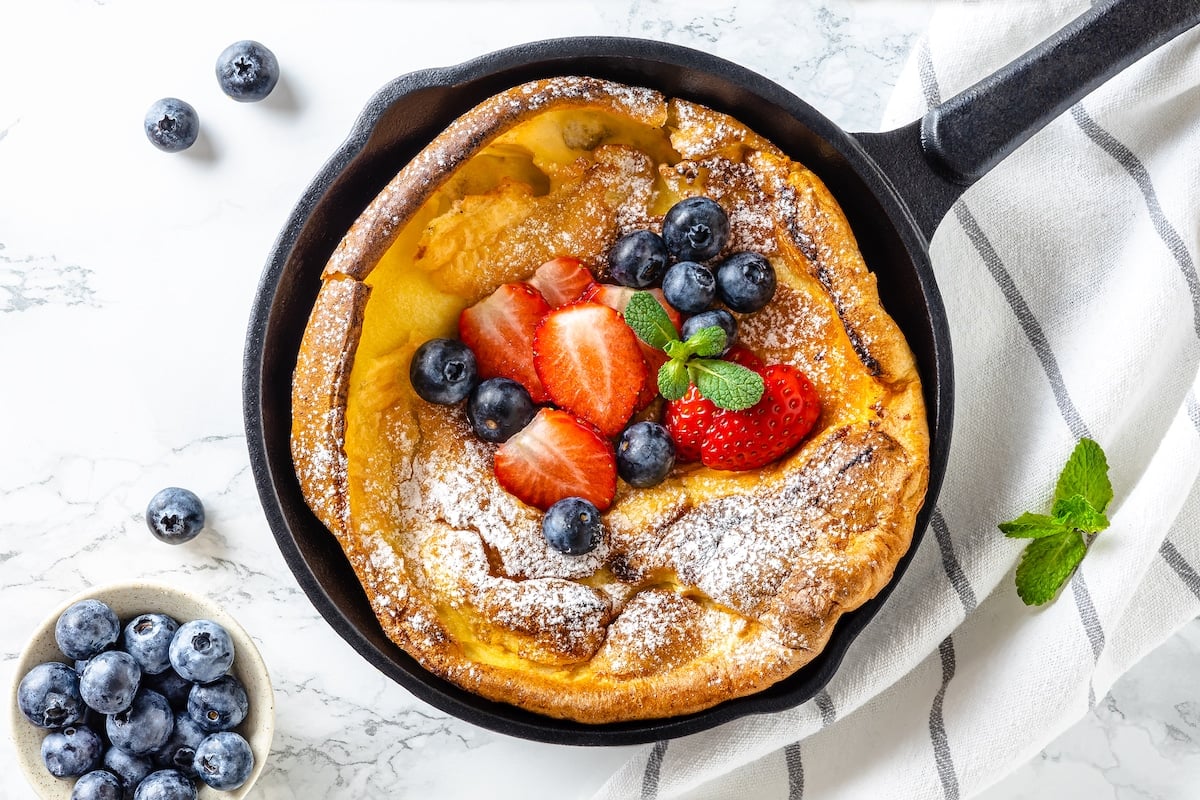 a cast iron skillet with a baked dutch baby and powdered sugar with fruits on top and a striped towel on the side.