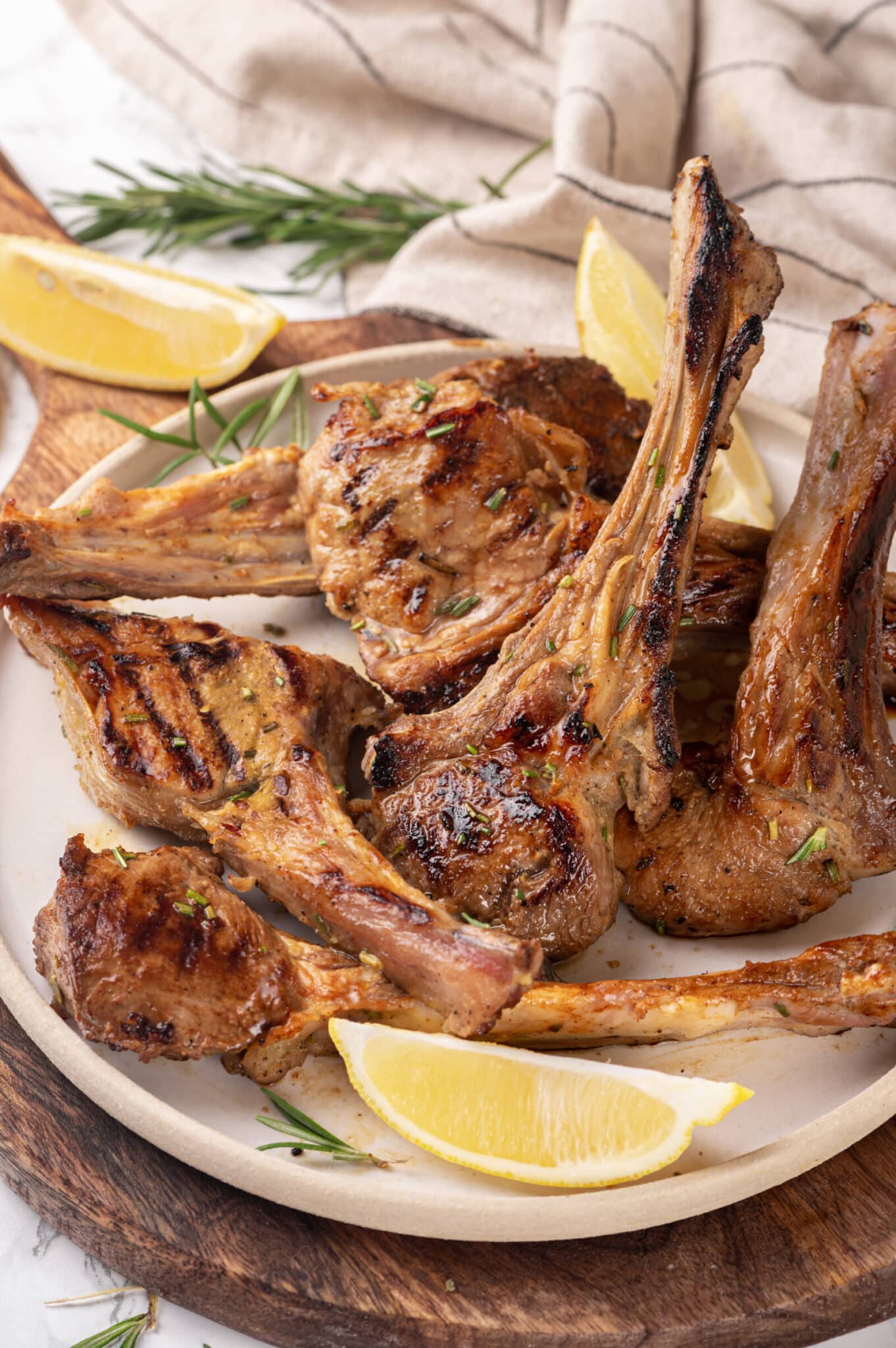 lamb chops on a beige plate and wood board with lemon wedges and rosemary.