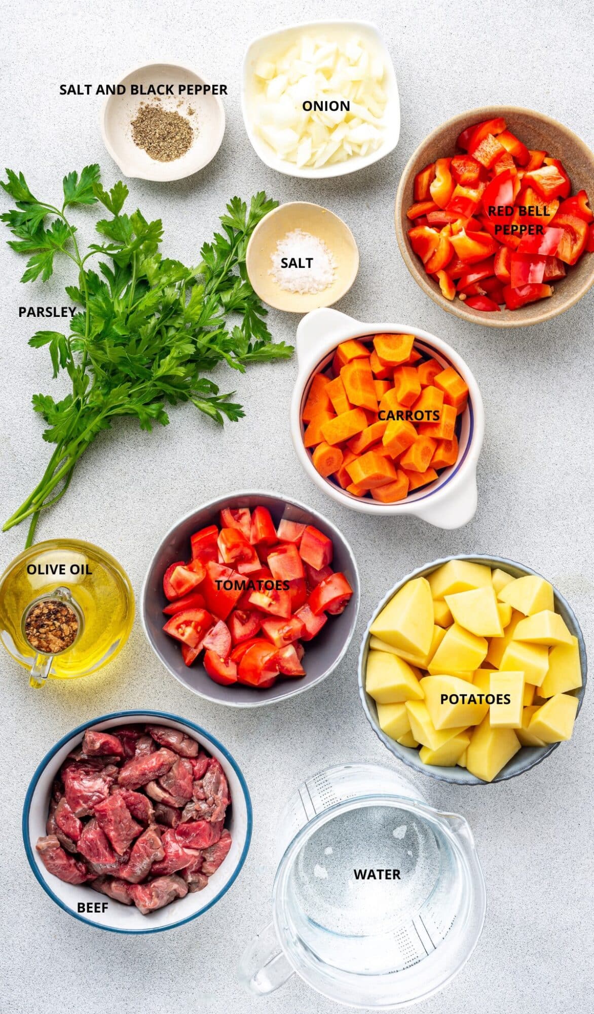 Chopped onion, red bell pepper, salt, carrots, potatoes, tomatoes, bottle of olive oil, parsley, cut up beef, and  measuring cup filled with water. 