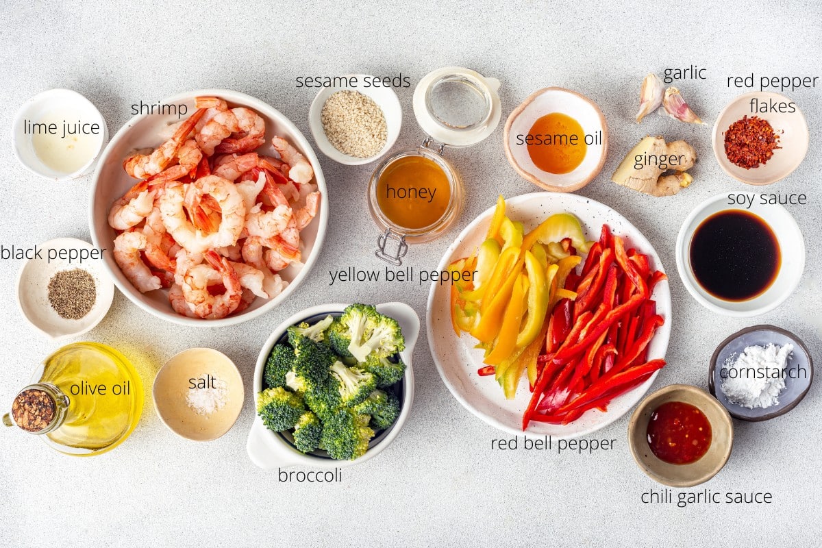 a spread of ingredients for a stir fry with labels corresponding to each ingredient in a thin black font lime juice shrimp sesame seeds honey sesame oil garlic ginger red pepper flakes soy sauce black pepper yellow bell pepper red bell pepper corn starch chili garlic sauce broccoli salt and olive oil.