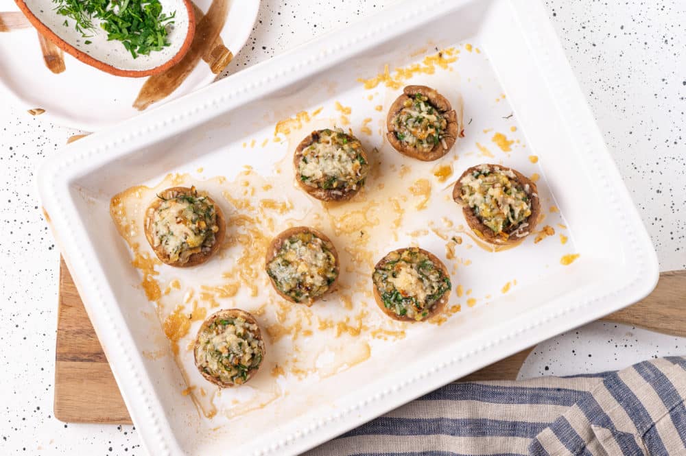 a wooden board with a white baking dish on it with prepared and baked stuffed mushroons.