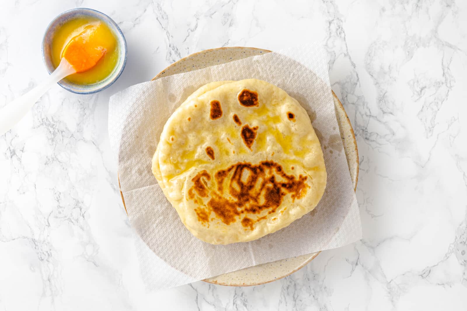 naan bread on a paper towel on a piece of plate with the melted butter to the left with a utensil to spread the butter in the bowl