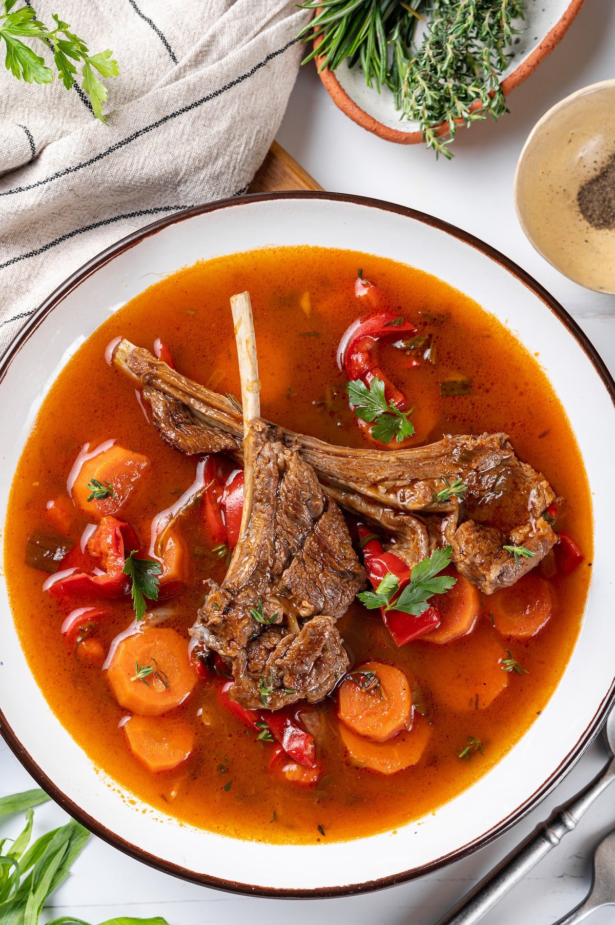 lamb chops in tomato water with herbs and chopped vegetables in a white bowl.