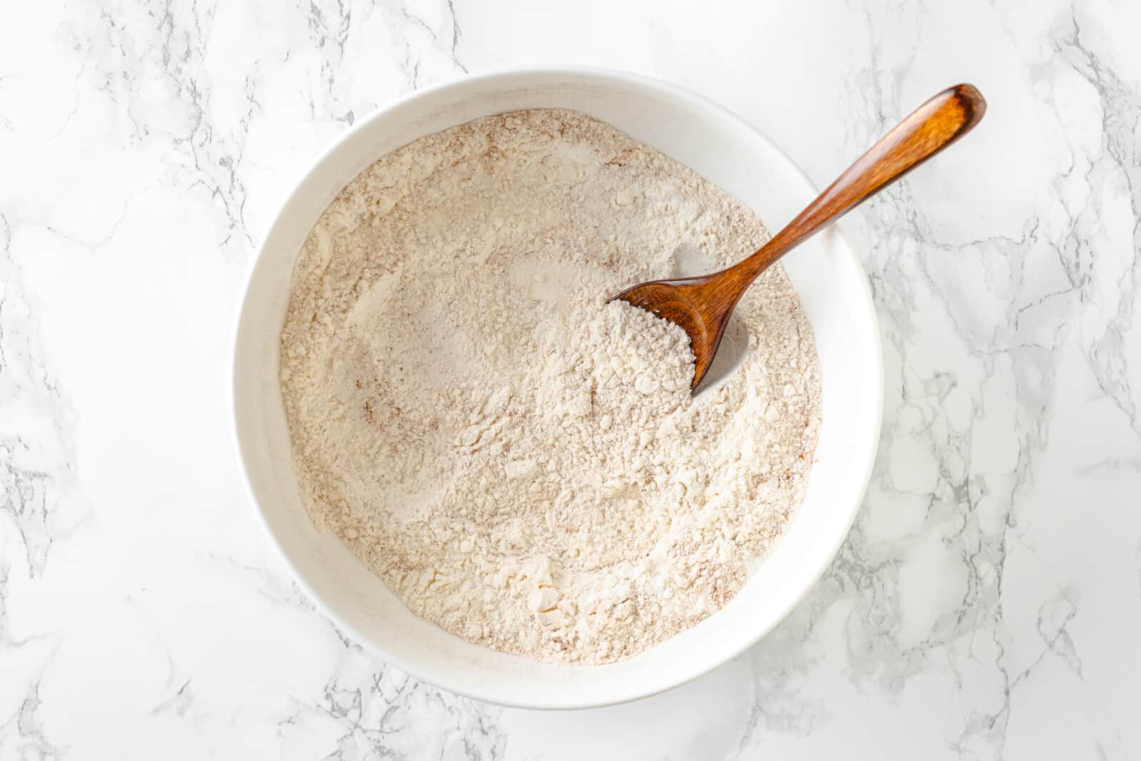 a white bowl of flour, sugar, salt, cinnamon, and baking powder mixed together with a wooden spoon