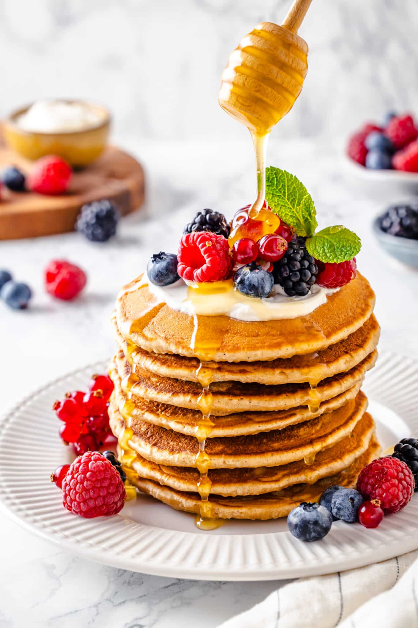 a large stack of thick pancaked with cream and berries on top and around it, with honey being drizzled over it 