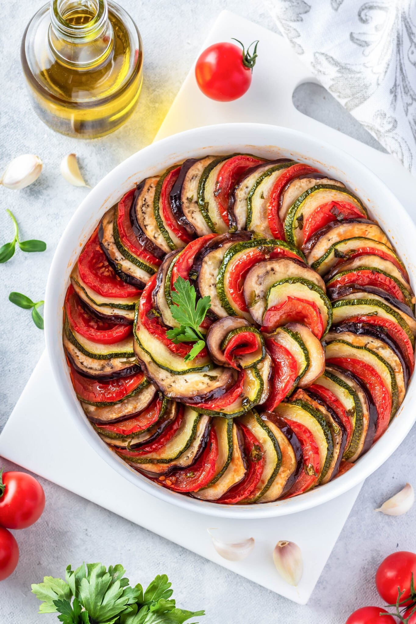 Ratatouille in a round casserole dish on a cutting board surrounded by ingredients for the dish.