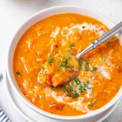 Red lentil soup in a bowl with a spoon, serving with cream and chopped greens for garnish.