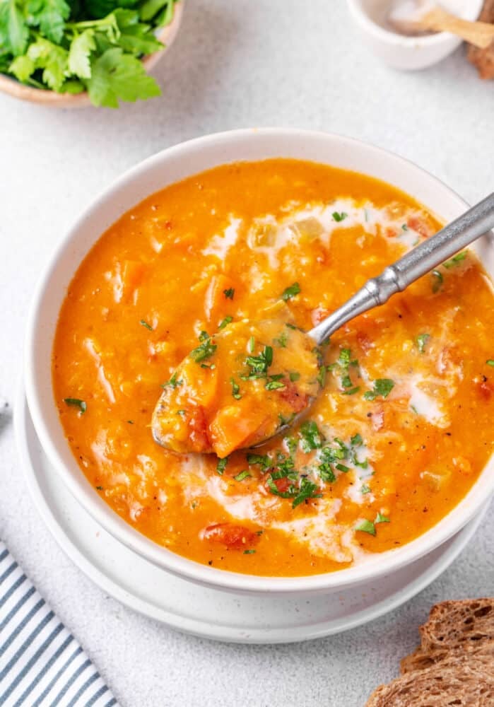 Red lentil soup in a bowl with a spoon, serving with cream and chopped greens for garnish.