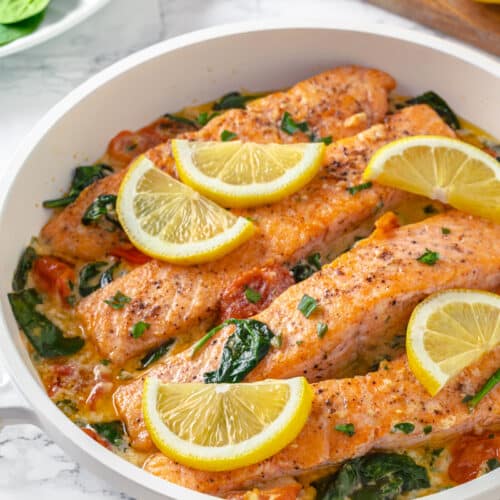 seared salmon filet in a creamy butter sauce with spinach and tomatoes in a white pan.