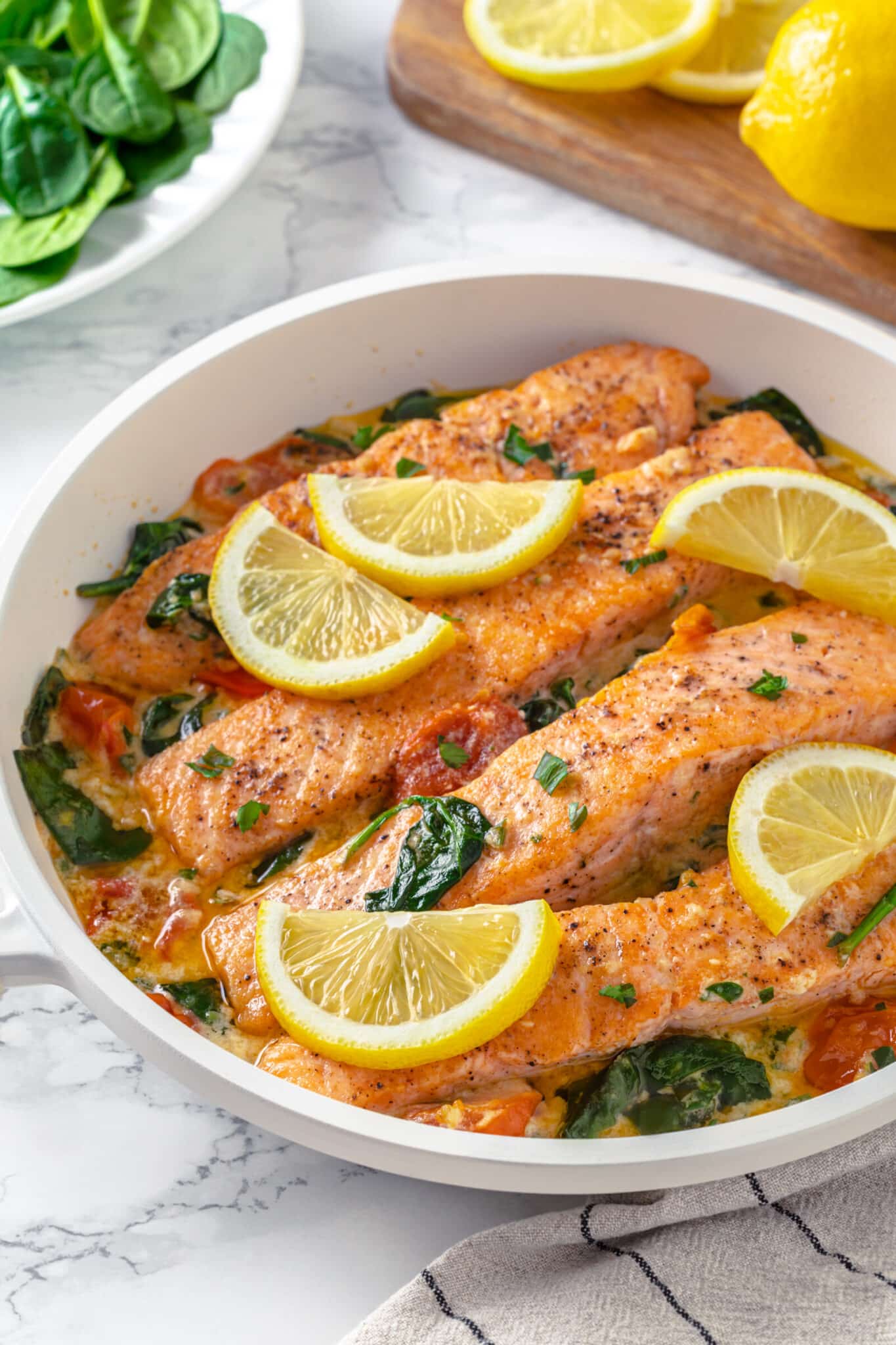 seared salmon filet in a creamy butter sauce with spinach and tomatoes in a white pan.