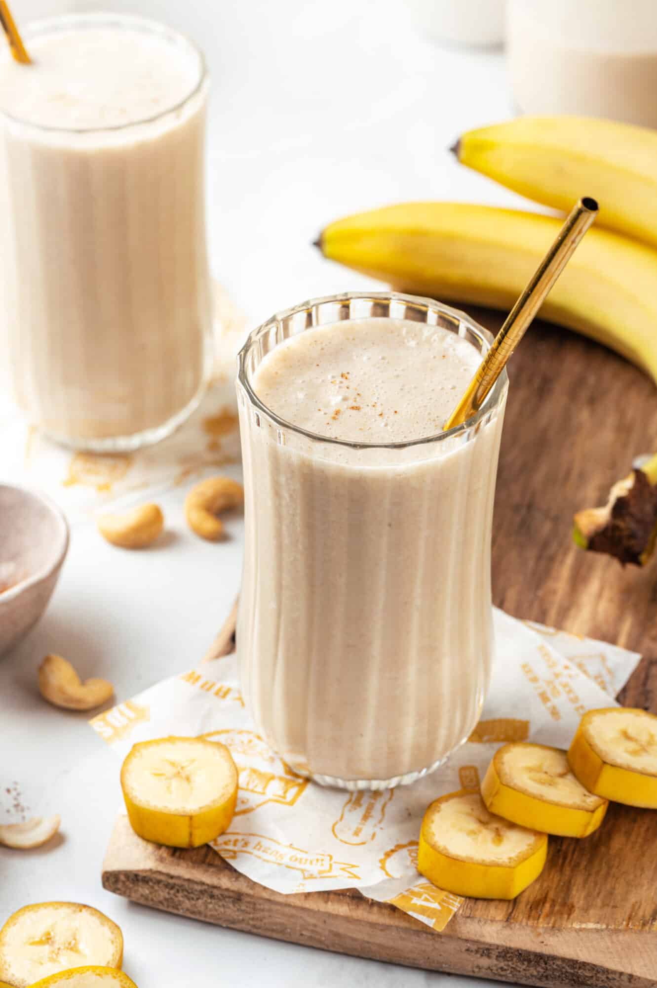 Serving banana bread smoothie in a glass atop a wooden board with banana slices.