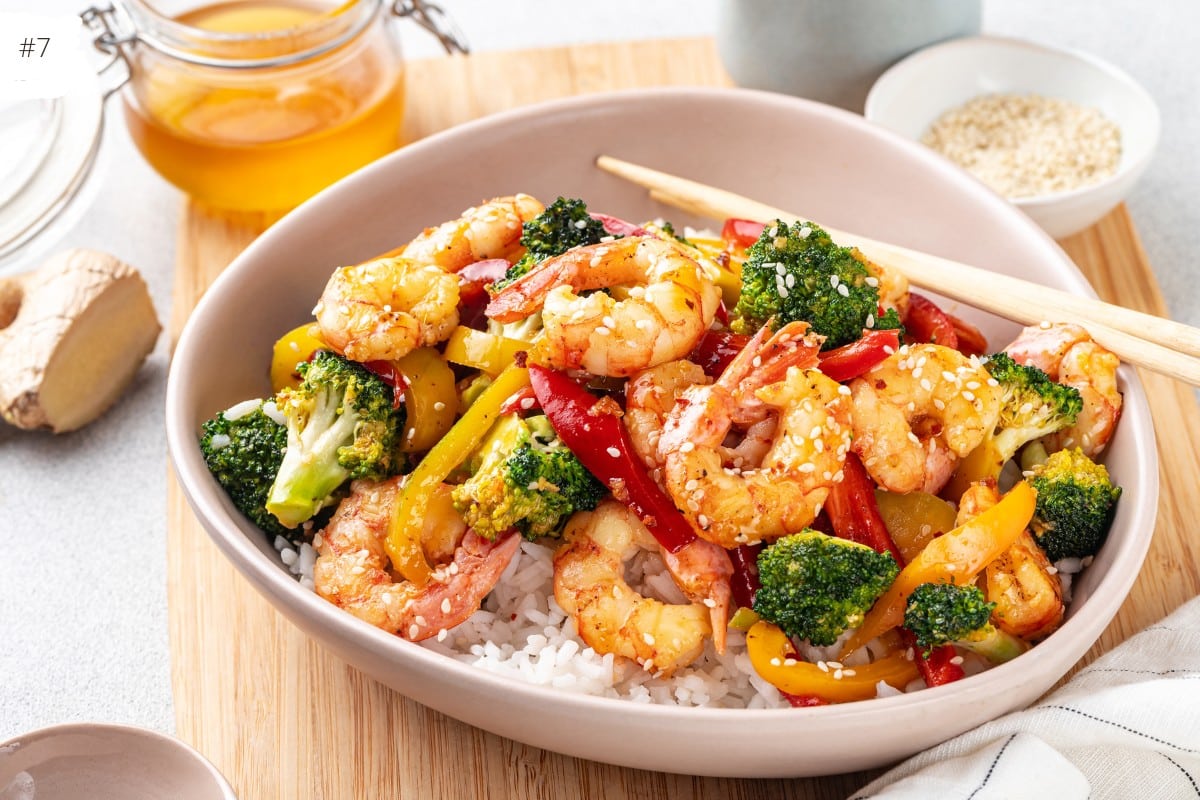 shrimp with broccoli and bell peppers in white shallow bowl over rice on wooden board.