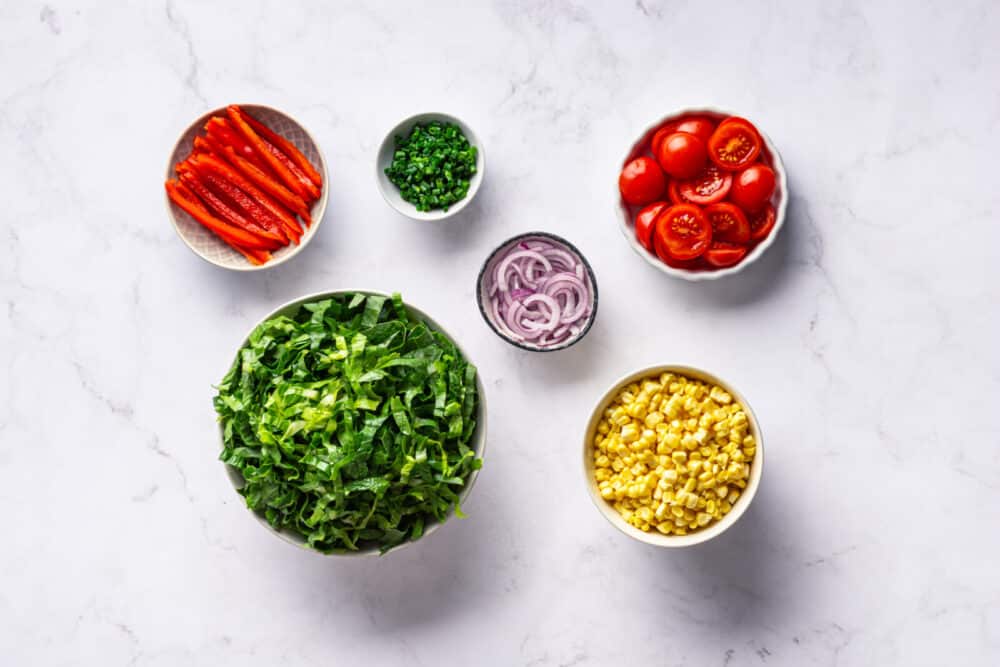 Smoked corn salad ingredients, prepared in bowls- chopped tomatoes, bell pepper, lettuce, chives, red onion, and corn.