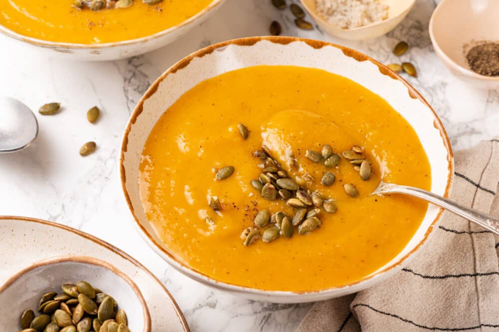 Squash soup in a bowl with pumpkin seeds on top, with a spoon for serving.