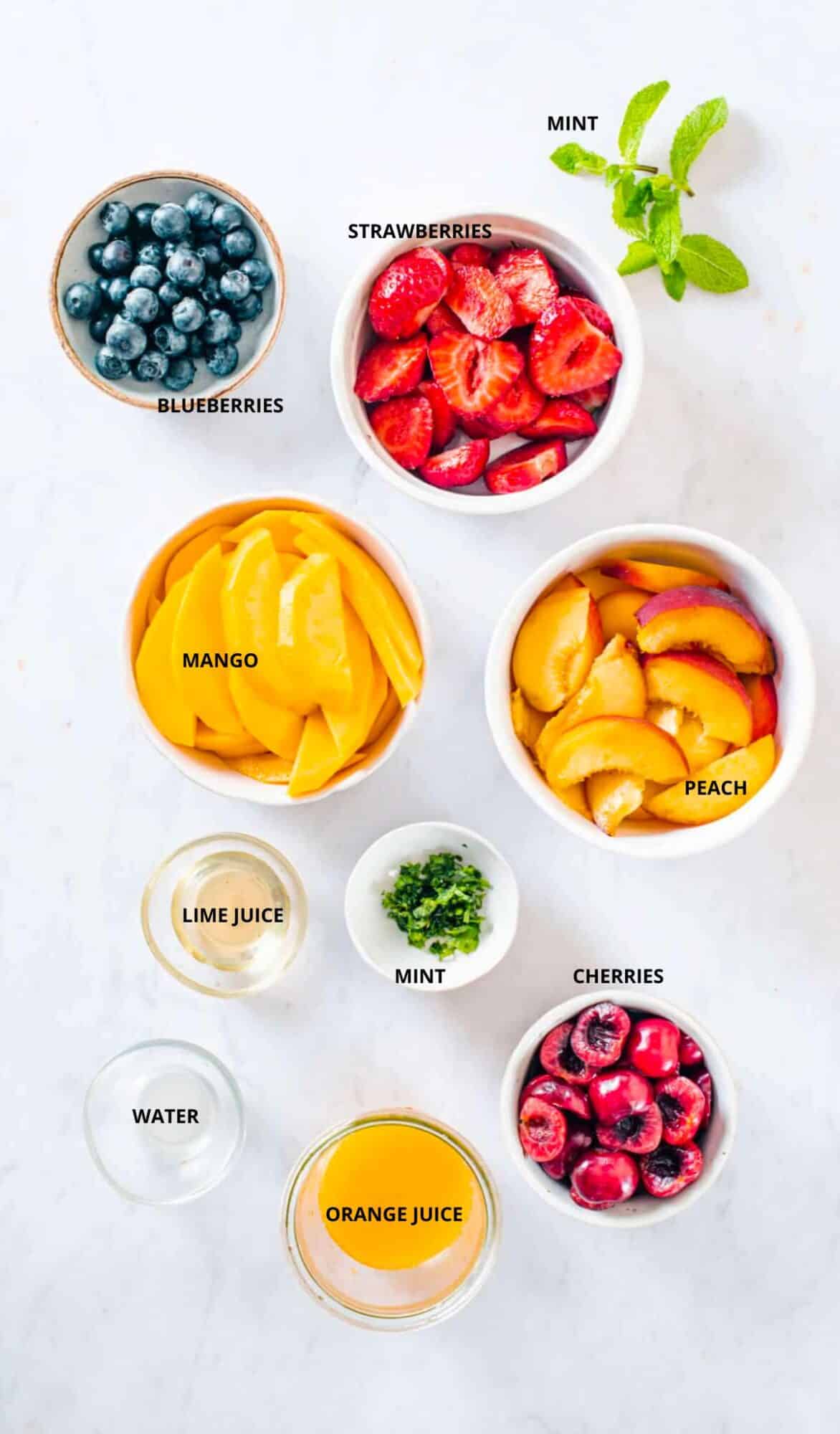 Summertime fruit salad ingredients with labels strawberries blueberries mint mango peaches lime juice cherries orange juice and water.