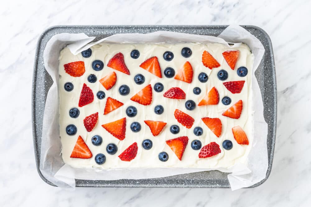 yogurt spread out in a pan with parchment paper with blueberries and sliced strawberries on top.