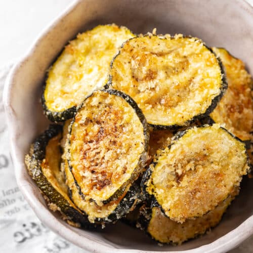 baked zucchini crisps in a white bowl on a newspaper.